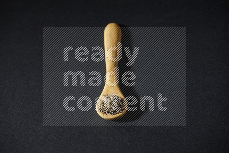 A wooden spoon full of cumin seeds on black flooring
