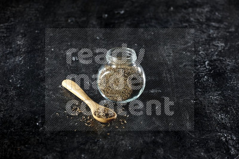 A glass spice jar and a wooden spoon full of cumin seeds on a textured black flooring