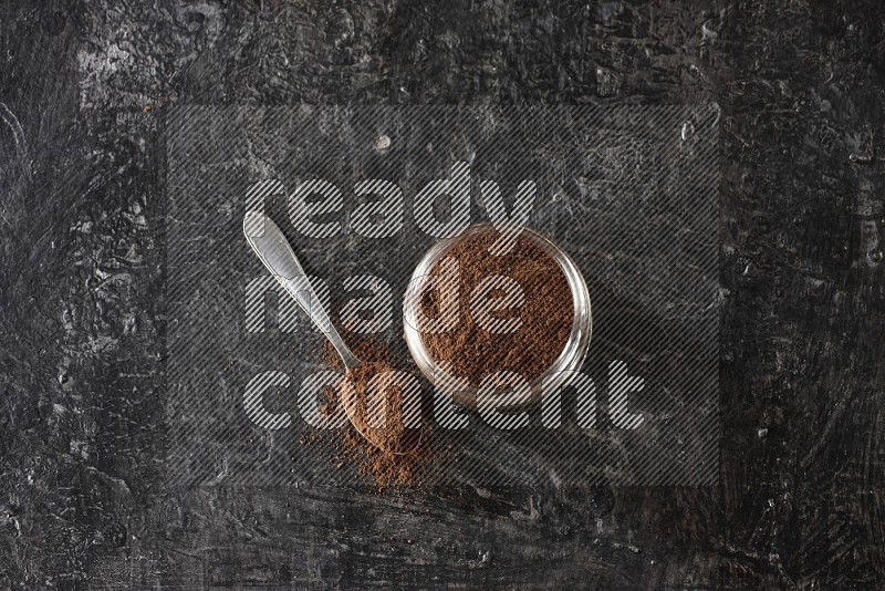 A glass jar full of cloves powder with a metal spoon on a textured black flooring