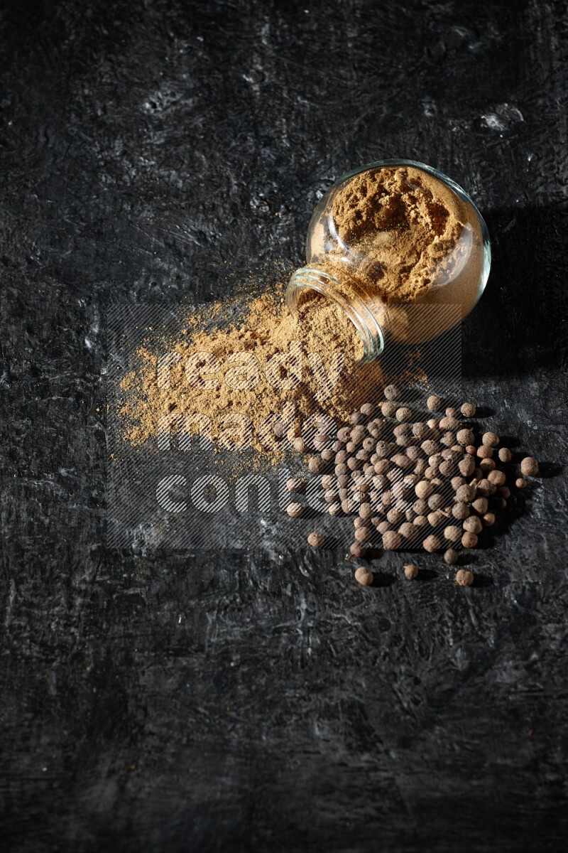 وعاء زجاجي للتوابل مقلوب ممتلئ ببودرة بهار الفلفل الحلو والبودرة تناثرت منه مع الحبوب على خلفية سوداء