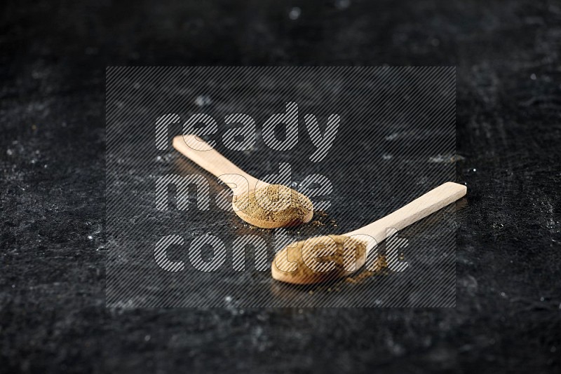 2 wooden spoons full of cumin powder on textured black flooring