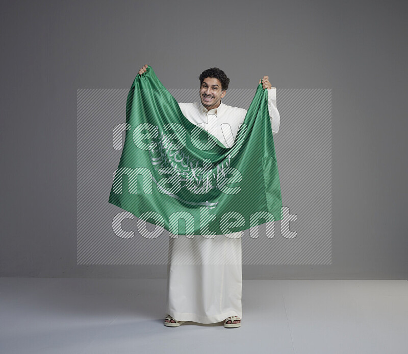 A Saudi man standing wearing thob holding big Saudi flag on gray background