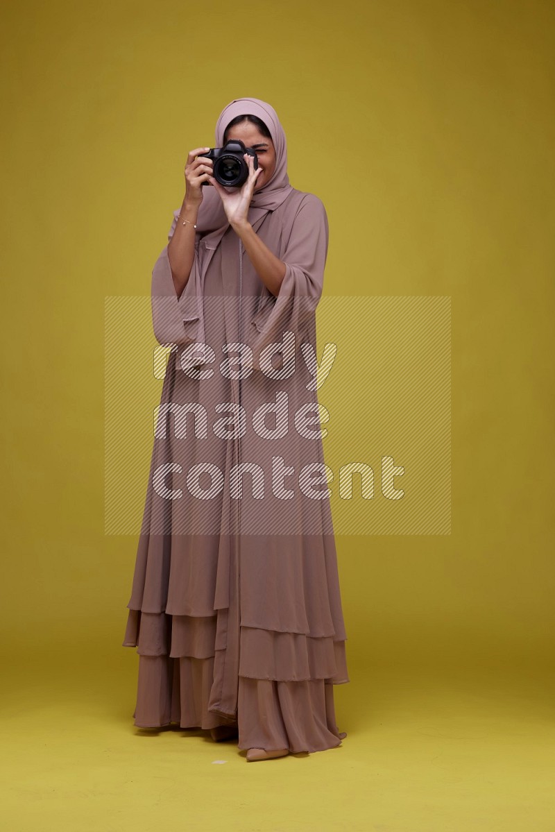 A woman Holding a Camera on a Yellow Background wearing Brown Abaya with Hijab