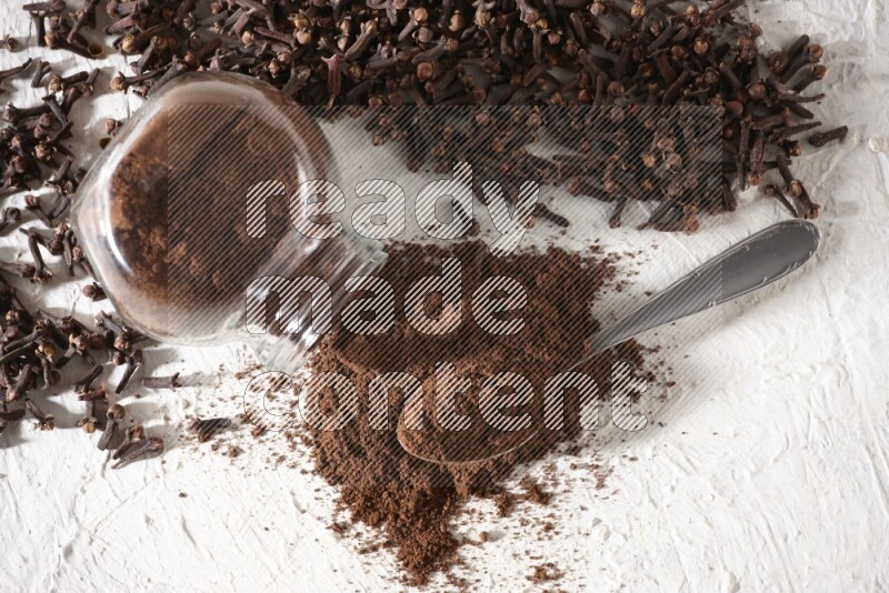 A flipped glass spice jar and a metal spoon full of cloves powder and powder came out of the jar with cloves spread on textured white flooring
