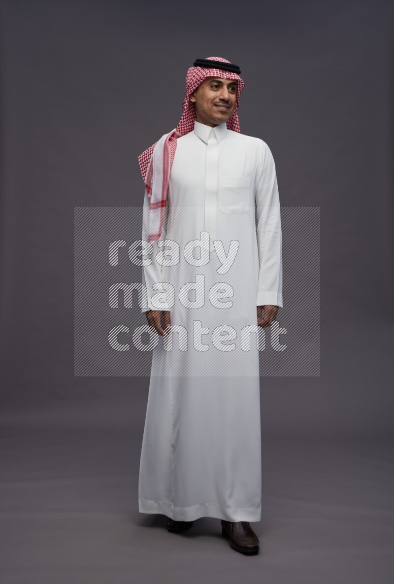 Saudi man wearing thob and shomag standing interacting with the camera on gray background