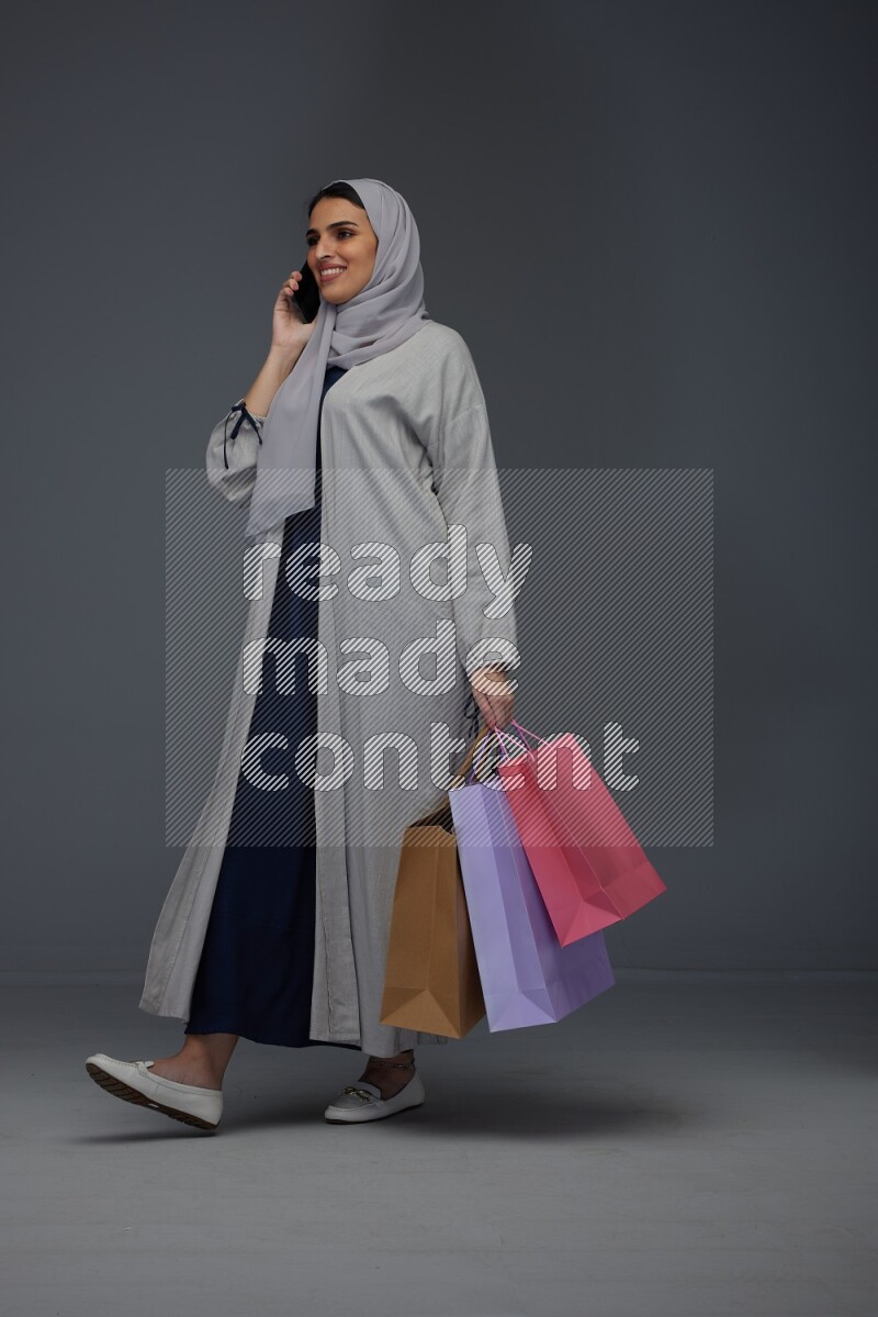 A Saudi woman wearing a light gray Abaya and head scarf standing and holding shopping bags making different poses eye level on a grey background