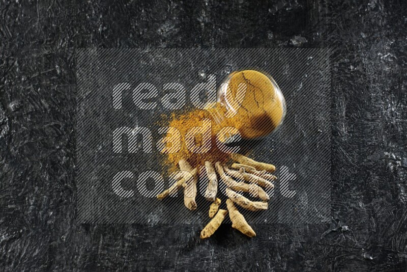 وعاء زجاجي للتوابل ممتلئ ببودرة الكركم مقلوب والبودرة تناثرت منه مع أصابع الكركم المجففة على خلفية سوداء