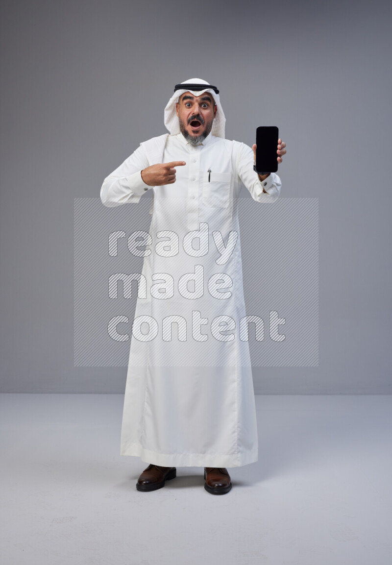 Saudi man Wearing Thob and white Shomag standing showing phone to camera on Gray background