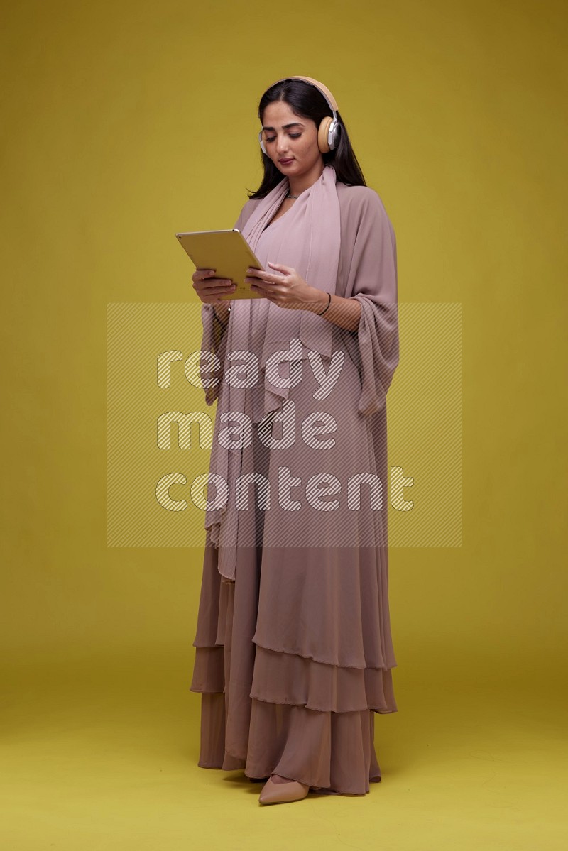 A woman Holding an Ipad with Headphone on a Yellow Background wearing Brown Abaya