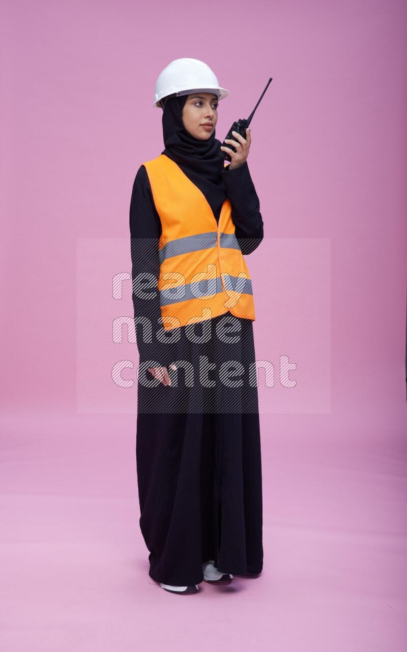Saudi woman wearing Abaya with engineer vest and helmet standing holding walkie-talkie on pink background