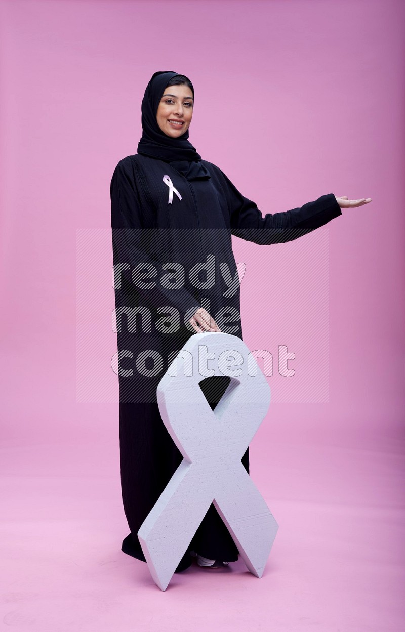 Saudi woman wearing pink ribbon on abaya standing holding awareness ribbon on plain pink background