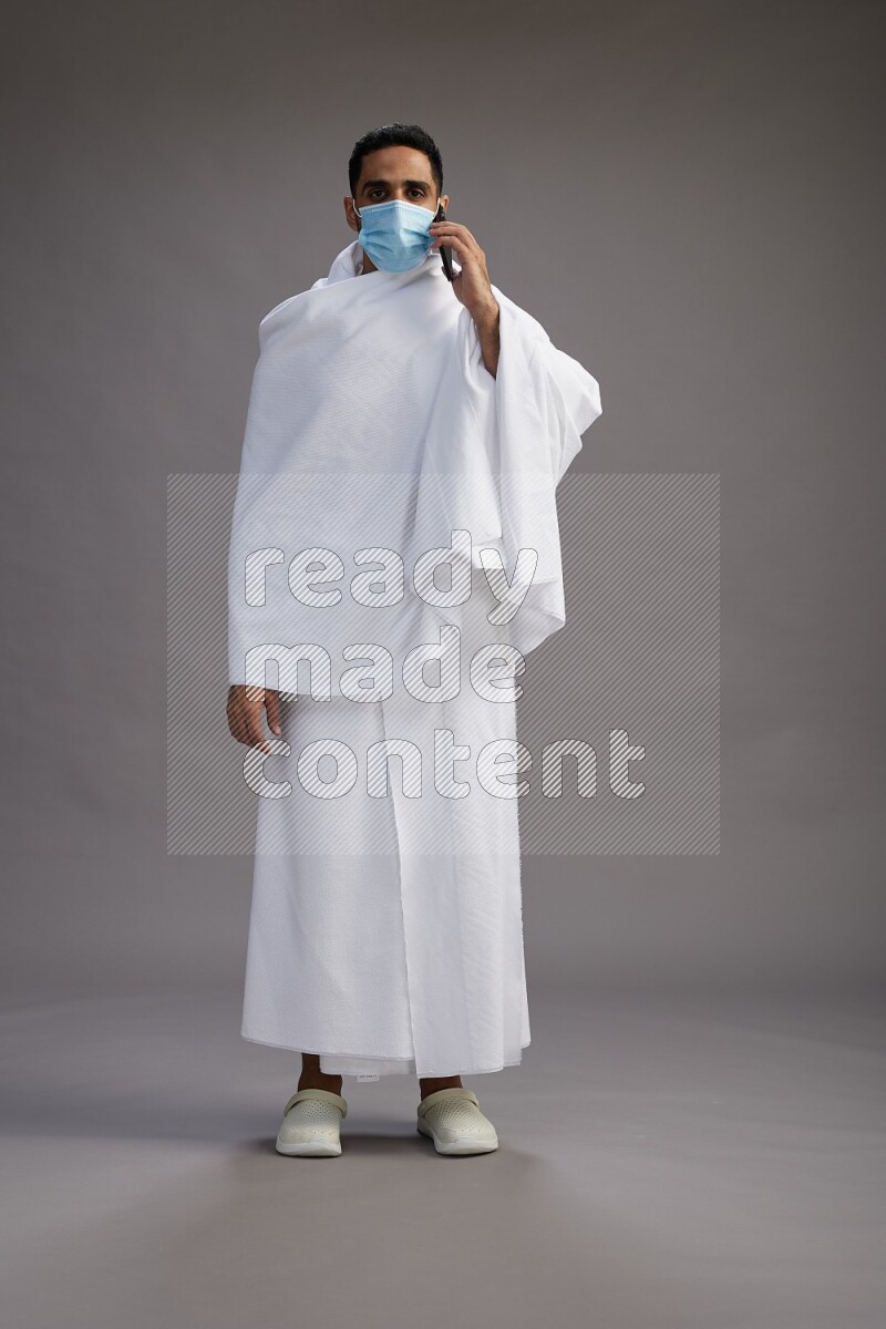A man wearing Ehram with face mask Standing talking on phone on gray background