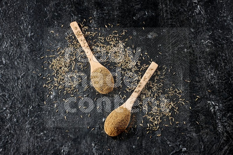 2 wooden spoons full of cumin powder with spreaded seeds on textured black flooring