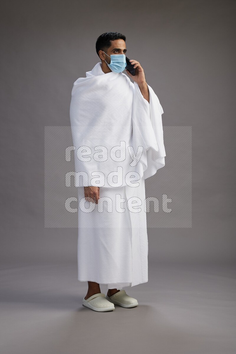 A man wearing Ehram with face mask Standing talking on phone on gray background