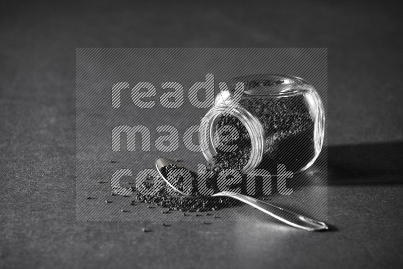 وعاء بهارات زجاجي مقلوب ممتلئ ببذور حبة البركة والبذور تناثرت منه مع ملعقة معدنية ممتلئة على أرضية سوداء