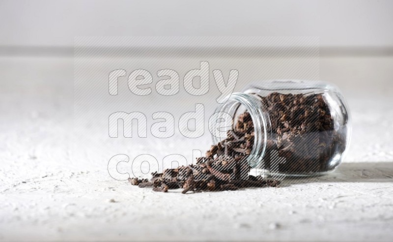 A flipped glass spice jar full of cloves and cloves came out of it on textured white flooring