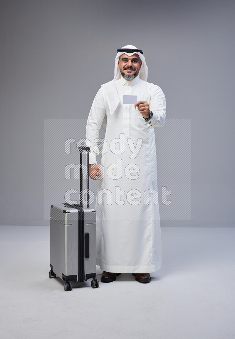 Saudi man wearing Thob and white Shomag standing holding Travel bag and ATM card on Gray background