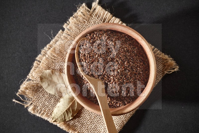 A wooden bowl full of flax with wooden spoon full of the seeds on it on burlap fabric on a black flooring in different angles