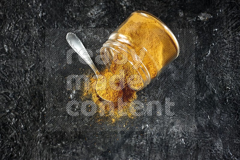 A flipped glass jar and a metal spoon full of turmeric powder and powder spilled out of it on textured black flooring