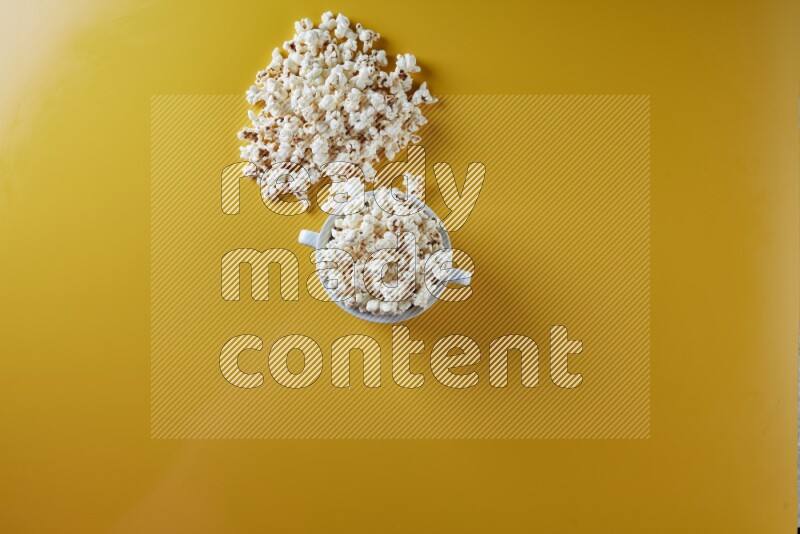 A white ceramic bowl full of popcorn with popcorn beside it on a yellow background in a top view shot