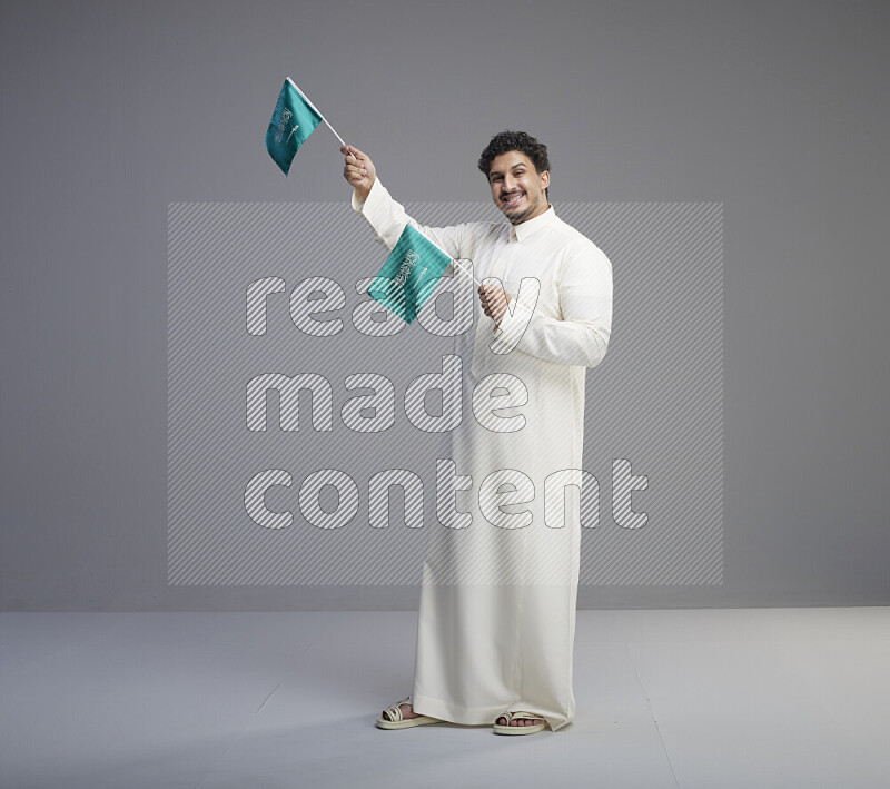رجل سعودي يرتدي ثوب ابيض يحمل علم السعودية على خلفية رمادي