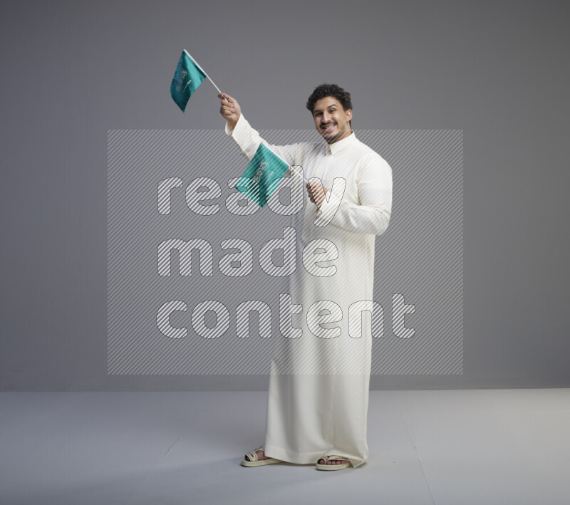 A Saudi man standing wearing thob holding small Saudi flag on gray background
