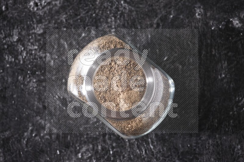 A glass spice jar full of black pepper powder on textured black flooring