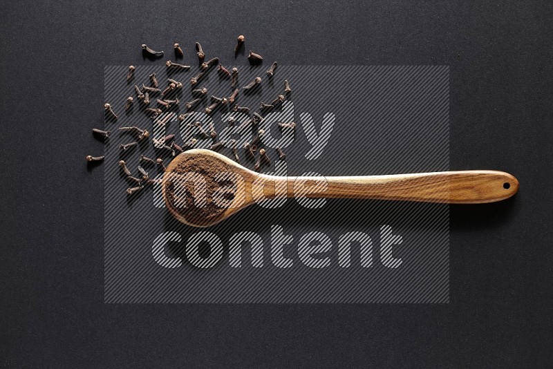 A wooden ladle full of cloves powder and some of whole cloves around it on a black flooring