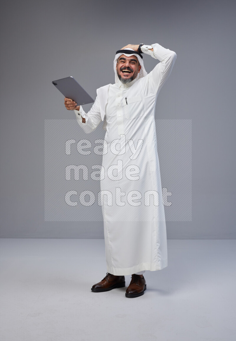 Saudi man Wearing Thob and white Shomag standing working on tablet on Gray background