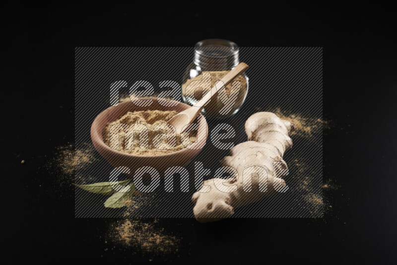 A wooden bowl full of ground ginger powder with a glass jar beside it and fresh ginger on black background