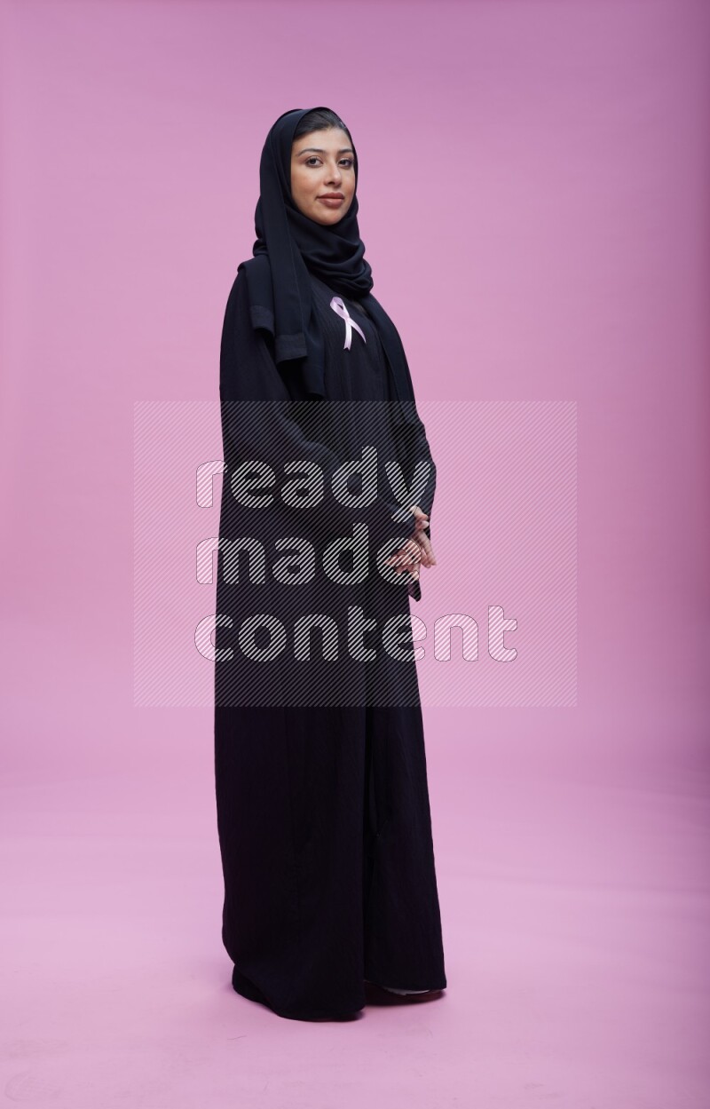 Saudi woman wearing pink ribbon on Abaya standing interacting with the camera on pink background