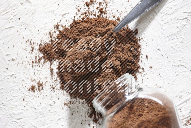 A flipped glass spice jar and a metal spoon full of cloves powder and powder came out of the jar on textured white flooring