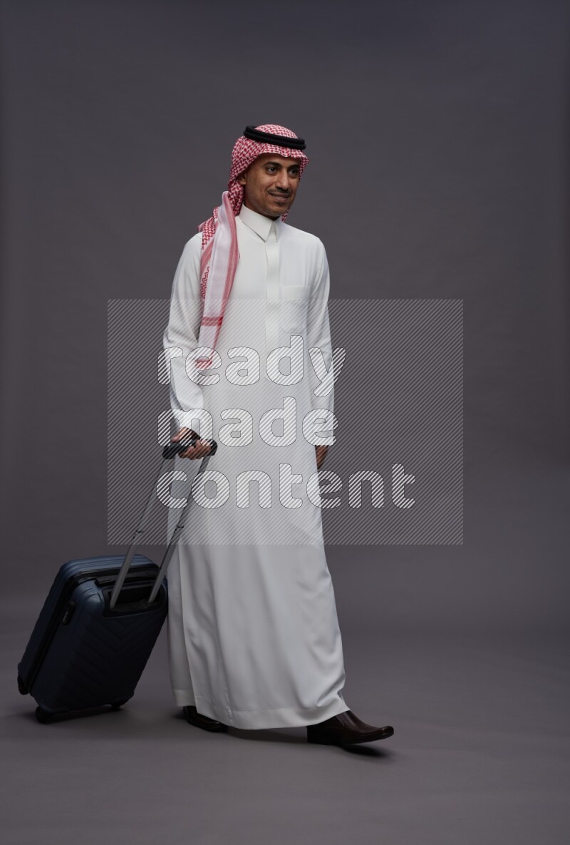 Saudi man wearing thob and shomag standing holding bag on gray background