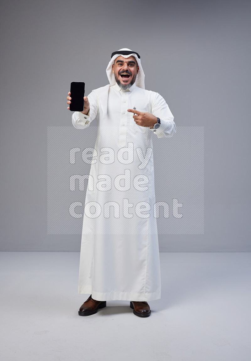 Saudi man Wearing Thob and white Shomag standing showing phone to camera on Gray background