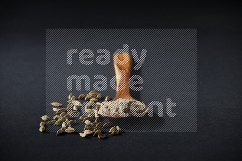 مغرفة خشبية ممتلئة ببودرة الهيل وحبوب الهيل بجانبها على خلفية سوداء