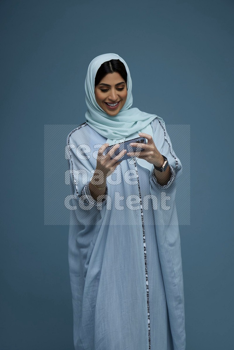 A Saudi woman Playing a Game on her phone on a blue background wearing a blue Abaya with hijab