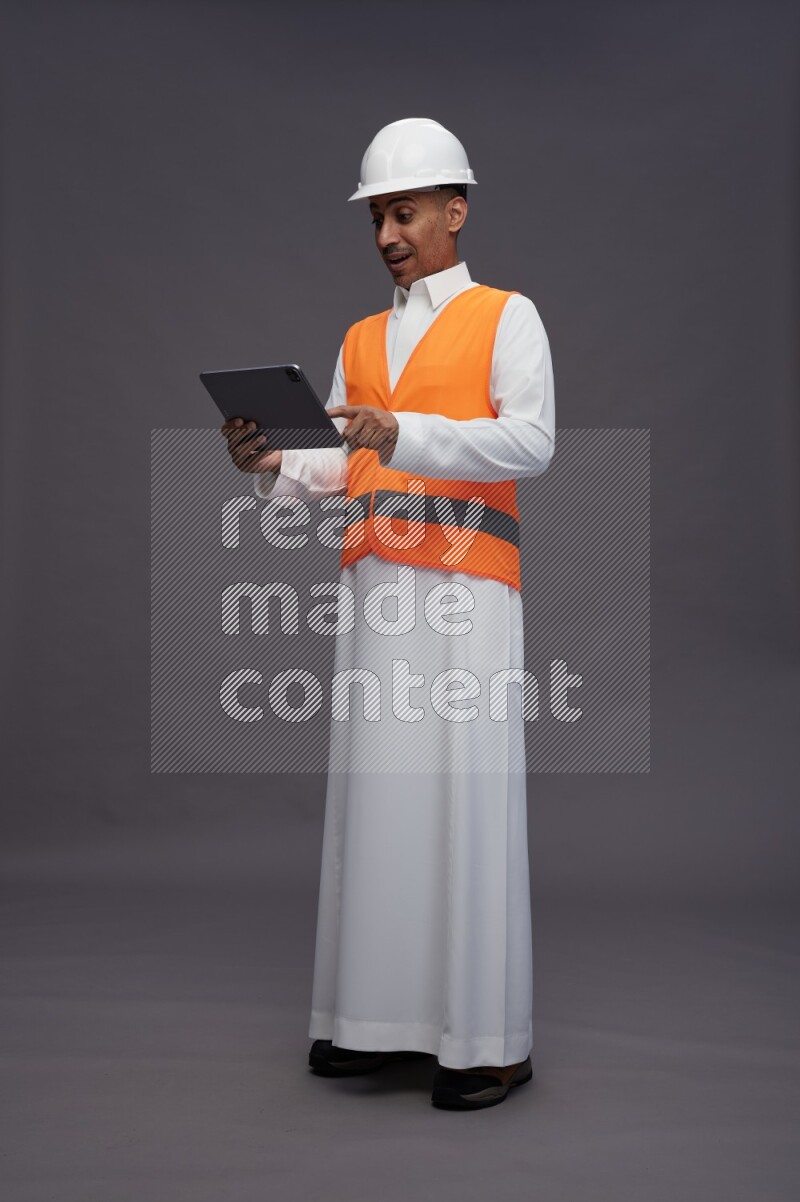 Saudi man wearing thob with engineer vest standing working on tablet on gray background