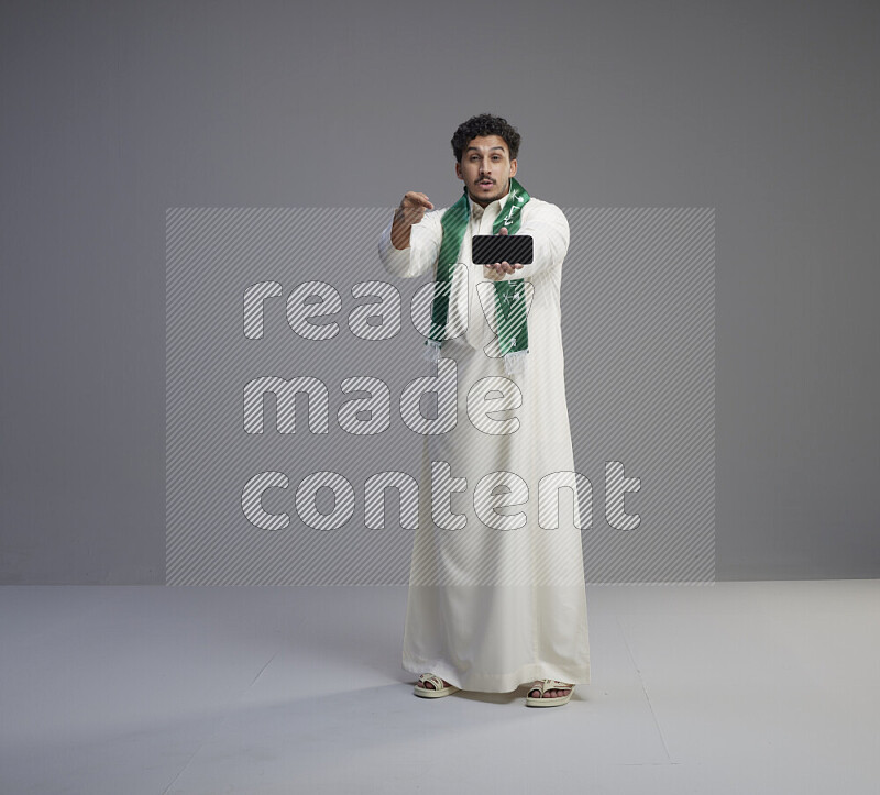 A Saudi man standing wearing thob and Saudi flag scarf showing phone to camera on gray background
