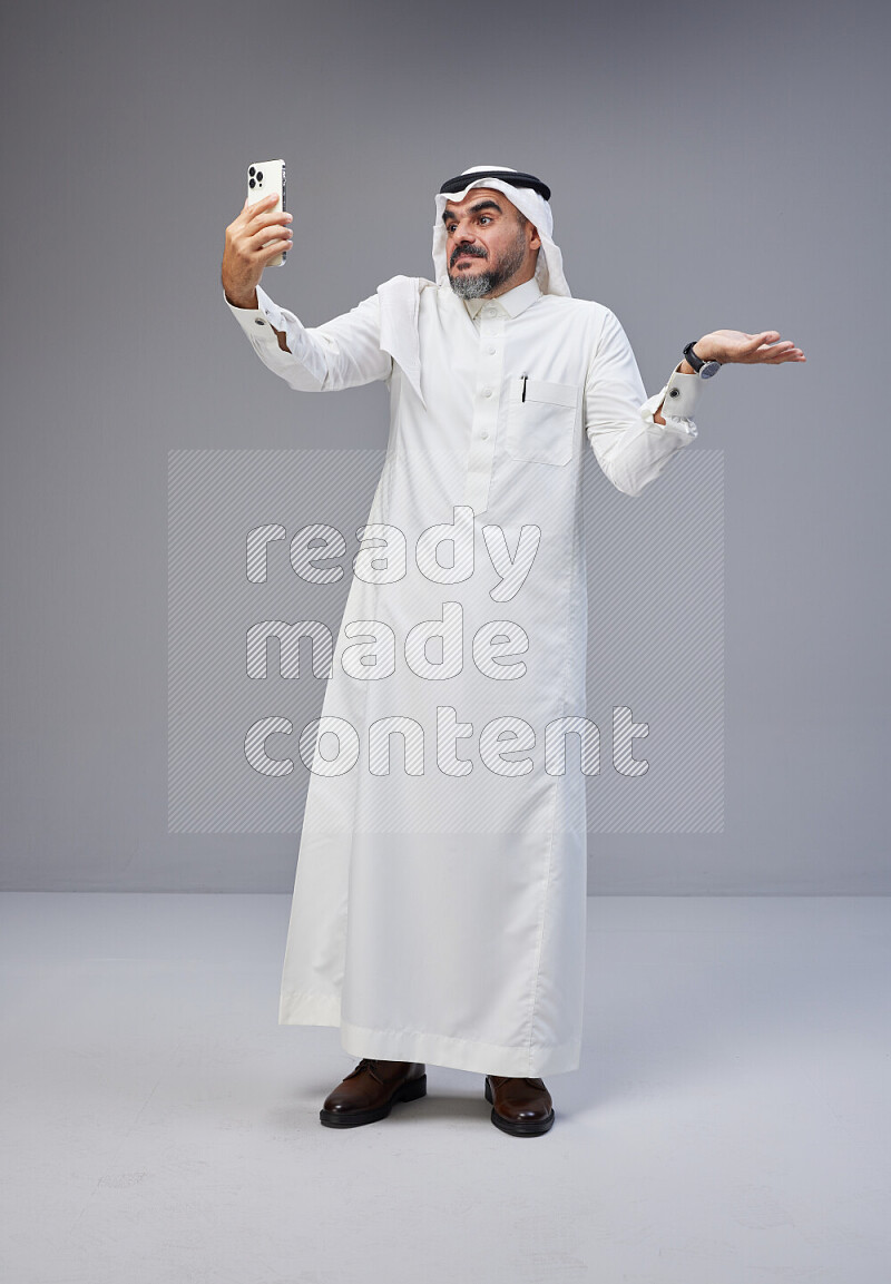 Saudi man Wearing Thob and white Shomag standing taking selfie on Gray background