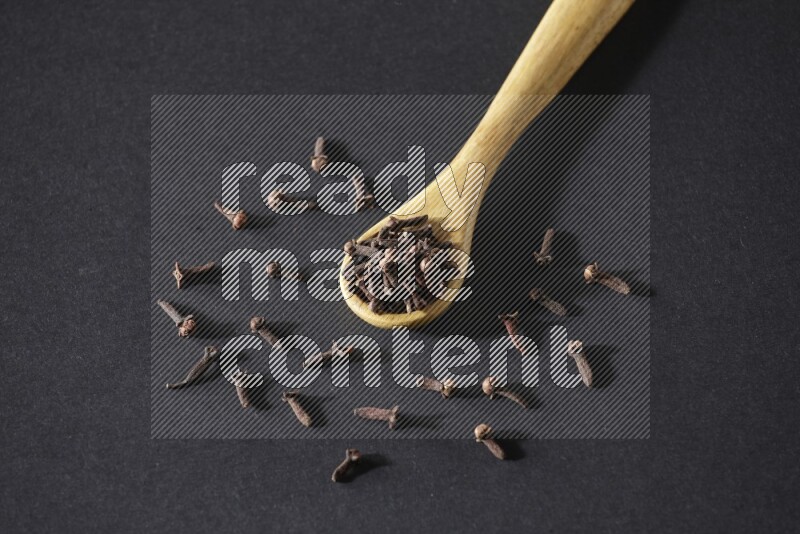 A wooden spoon full of cloves and some spreaded out of it on a black flooring