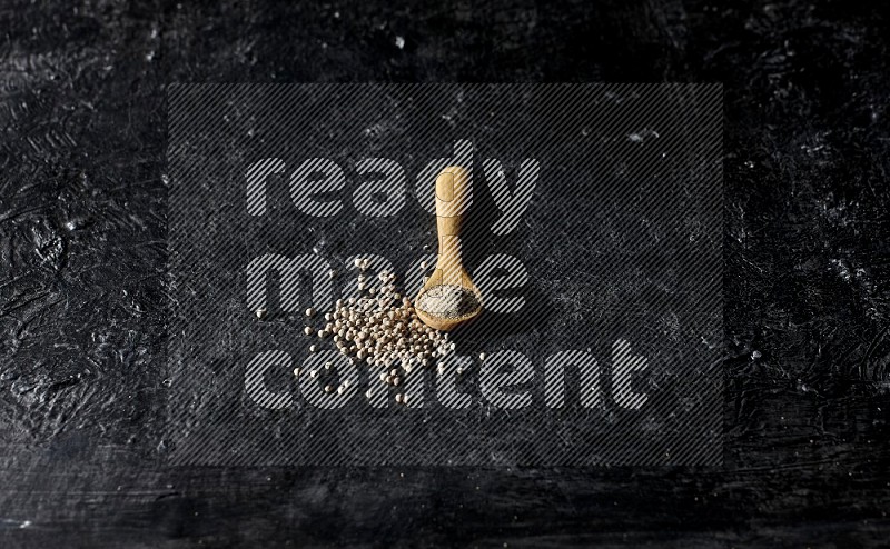 A wooden spoon full of white pepper powder with white pepper beads on textured black flooring