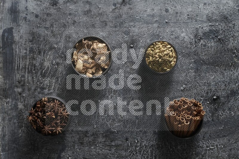 Ginger, Cardamom, Star anise and cinnamon sticks in 4 bowls on a textured black background