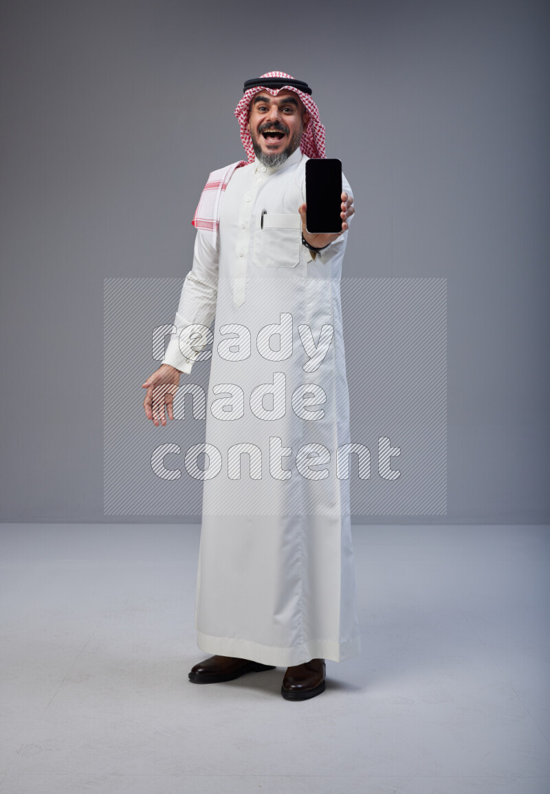 Saudi man Wearing Thob and red Shomag standing showing phone to camera on Gray background