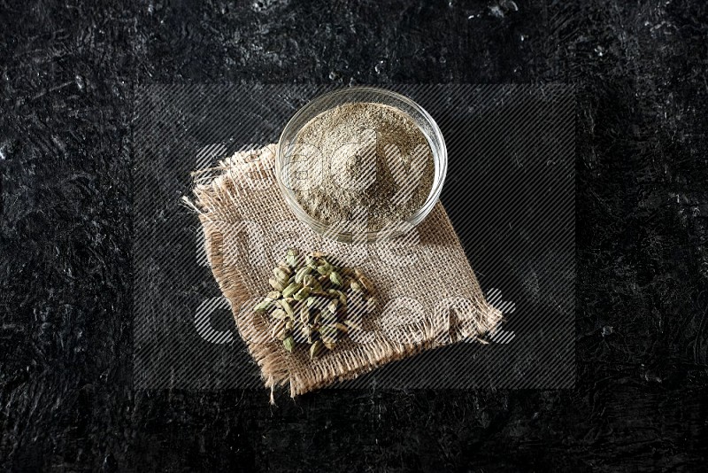 A glass bowl full of cardamom powder with cardamom seeds on a burlap piece on textured black flooring