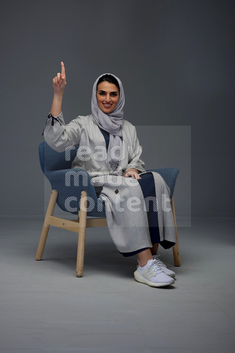 A Saudi woman wearing a light gray Abaya and head scarf sitting on a dark grey chair while making multi hand gestures eye level on a grey background