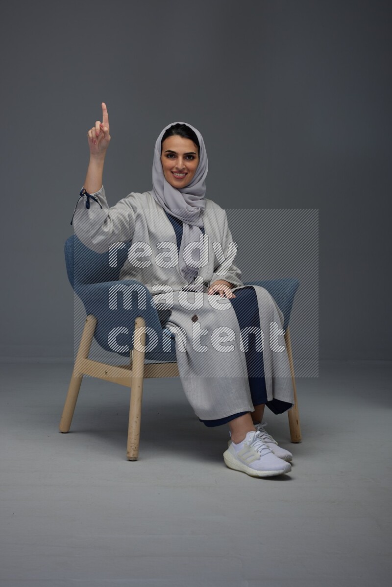 A Saudi woman wearing a light gray Abaya and head scarf sitting on a dark grey chair while making multi hand gestures eye level on a grey background