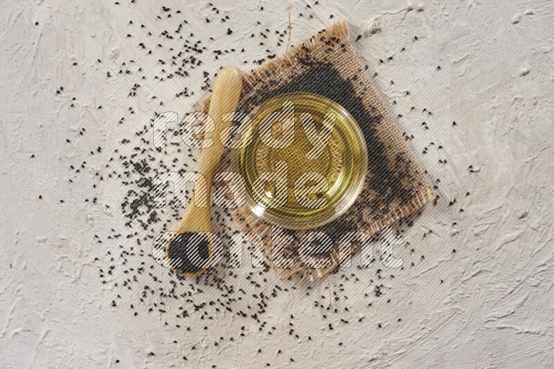 A glass bowl full of black seeds oil and wooden spoon full of black seeds with seeds spread on burlap fabric on a textured white flooring