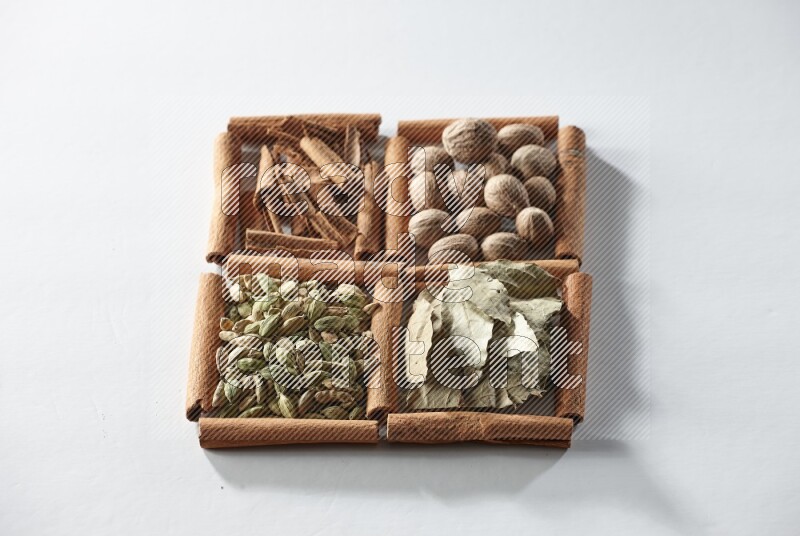 4 مربعات من أعواد القرفة ممتلئة بجوزة الطيب وأوراق الغار وحبوب الهيل والقرفة على أرضية بيضاء