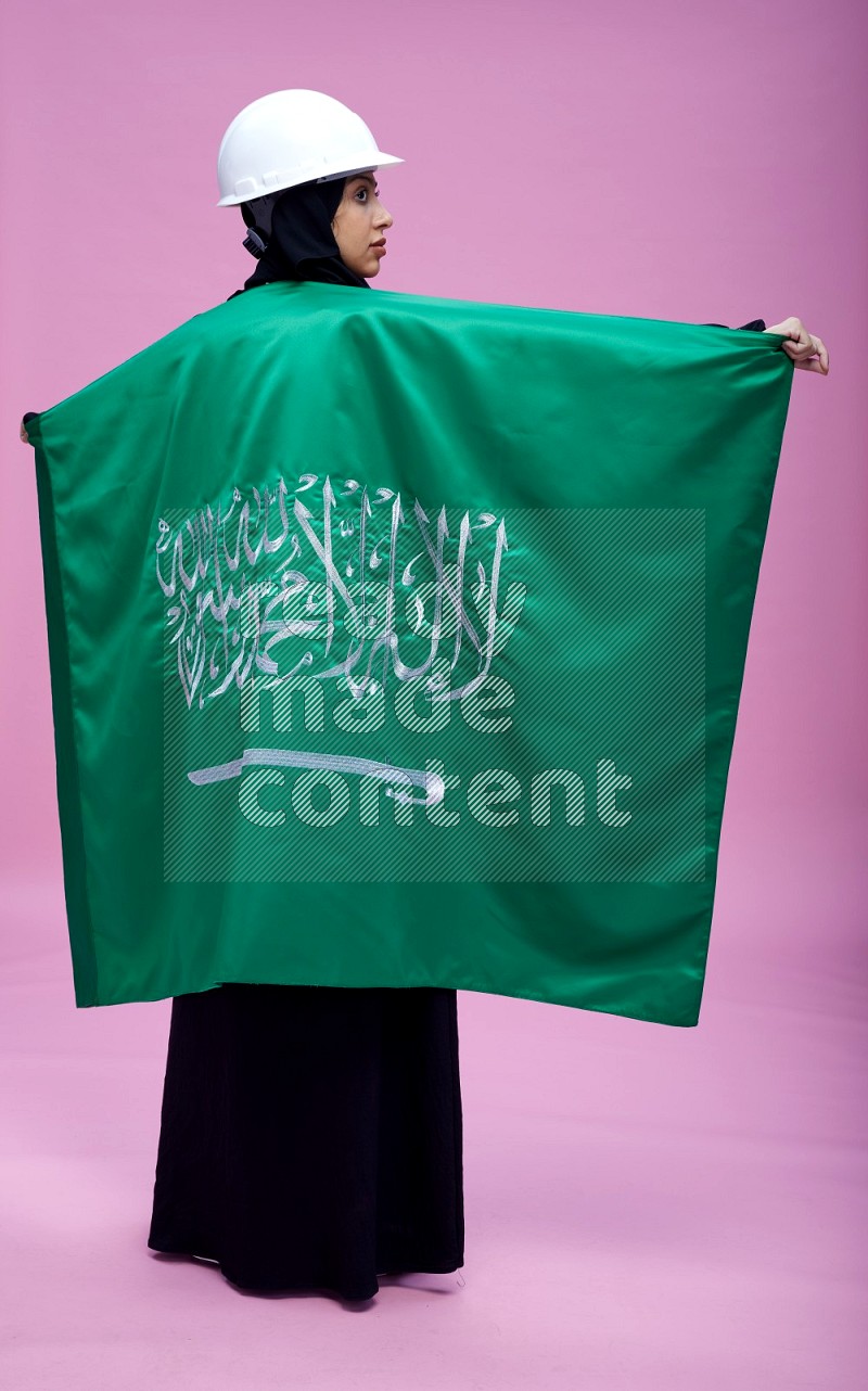 Saudi woman wearing Abaya with engineer vest and helmet standing holding Saudi flag on pink background