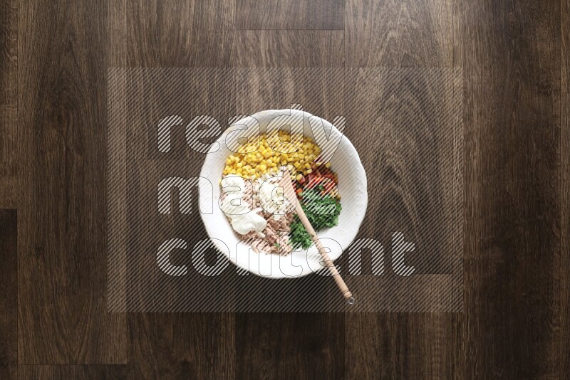 A white bowl full of tuna, colored bell pepper, sour cream, corn, parsley, black pepper powder and sauce, with wooden spoon on wooden background